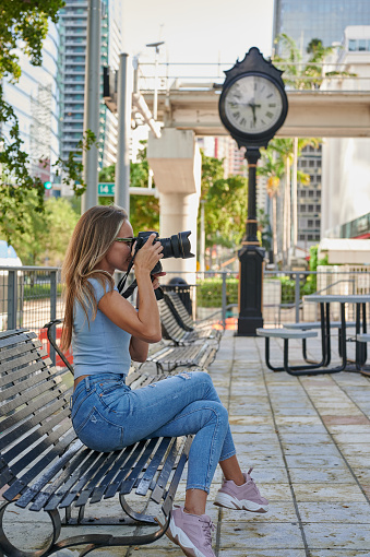 Female photographer photographing with DSLR camera sitting on the bench empty street