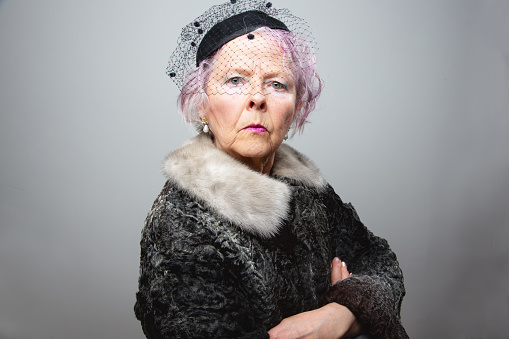 senior woman, wearing black hat with veil, fur coat, pearl ear rings and pink hair, arms crossed, looking into to camera with serious expression
