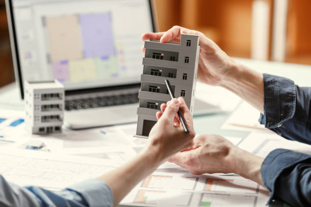 istock Two architects holding and discussing model of a house and architectural plans in a studio with blueprints and laptop on a table. 1220385273