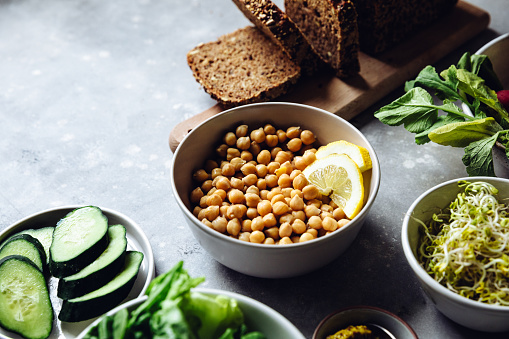 Close-up of bowl of chickpeas with lemon slices with sourdough bread, cucumber, and sprouts on table for preparing vegetarian sandwich for lunch.