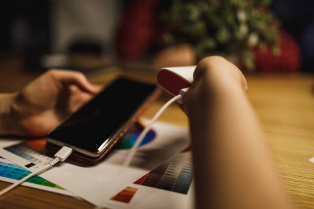 Close up of a female hand holding a portable battery pack that is connected to the smartphone on the table with several papers placed under and on top of the table.