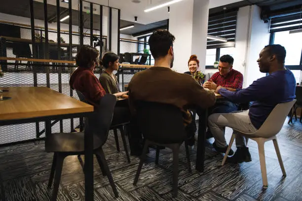 Wide shot of a group of office coworkers sitting at the table in the lounge area and sharing enjoyable time together.