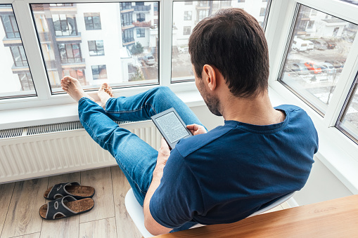 Relaxing man in a blue jeans and t-shirt reading an electronic book on digital device on the balcony by the window putting bare feet on a window sill in a bright modern apartment