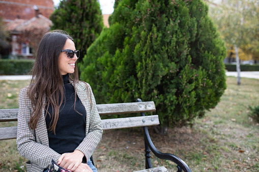 A young woman is sitting on a park bench and looking at something in the distance smiling