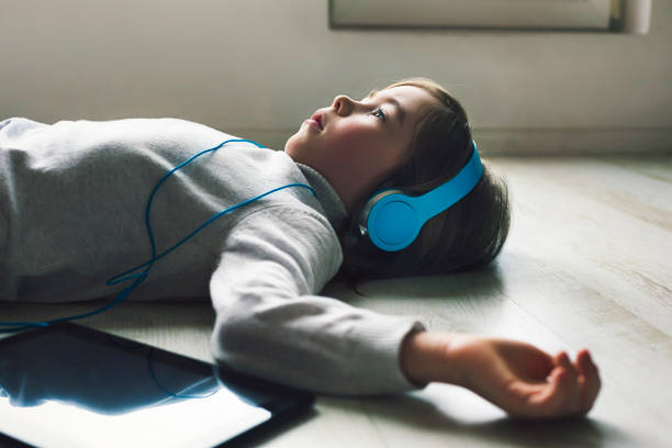 Sad child with blue headphones and digital tablet listening music at home. Lying on the floor stock photo
