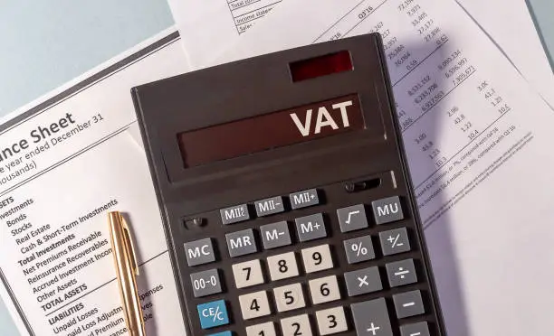 Photo of VAT word on display of calculator on papers documents and golden pen