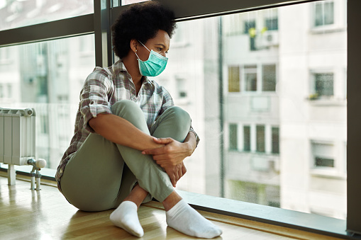 Pensive black woman with face mask looking through the window while sitting on the floor at home.