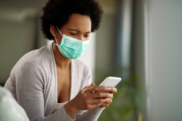 istock Black woman wearing face mask while text messaging on mobile phone at home. 1220416235