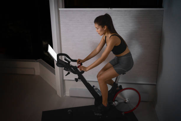 entrenamiento en casa en el interior bicicleta estática asiática chica ciclismo pantalla con clases en línea mujer entrenamiento en equipo de fitness inteligente en interiores para el ejercicio de ciclismo. tarde en la noche en el dormitorio - bicicleta estática fotografías e imágenes de stock