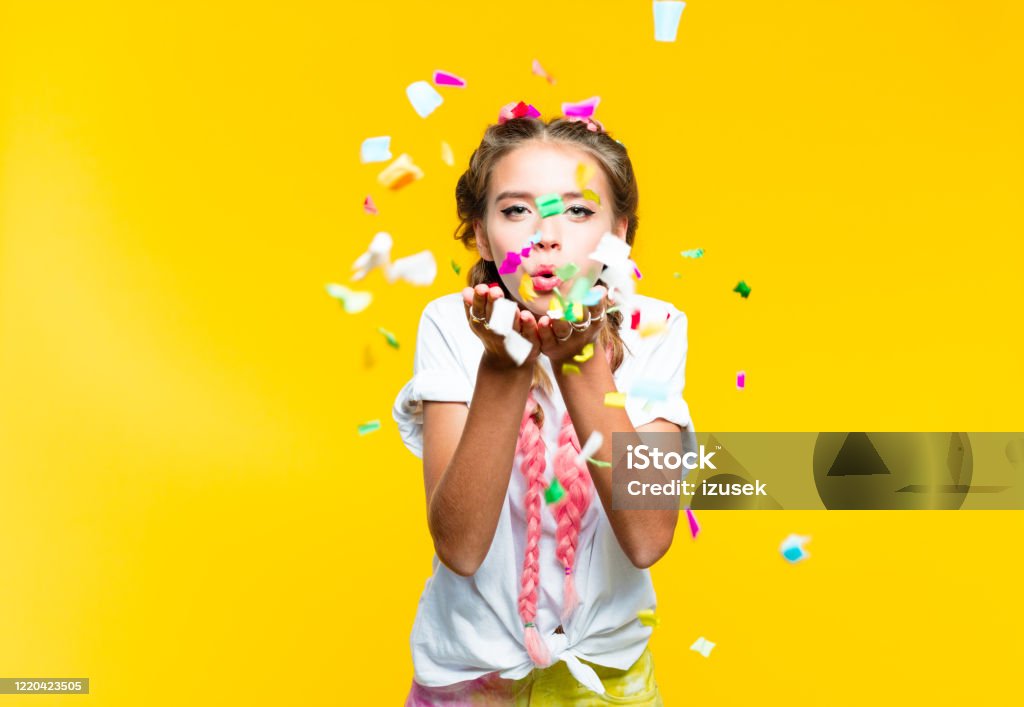 Teenage girl blowing colorful confetti Happy teenage girl with pink braids wearing white t-shirt blowing colorful confetti. Studio shot, yellow background. Confetti Stock Photo
