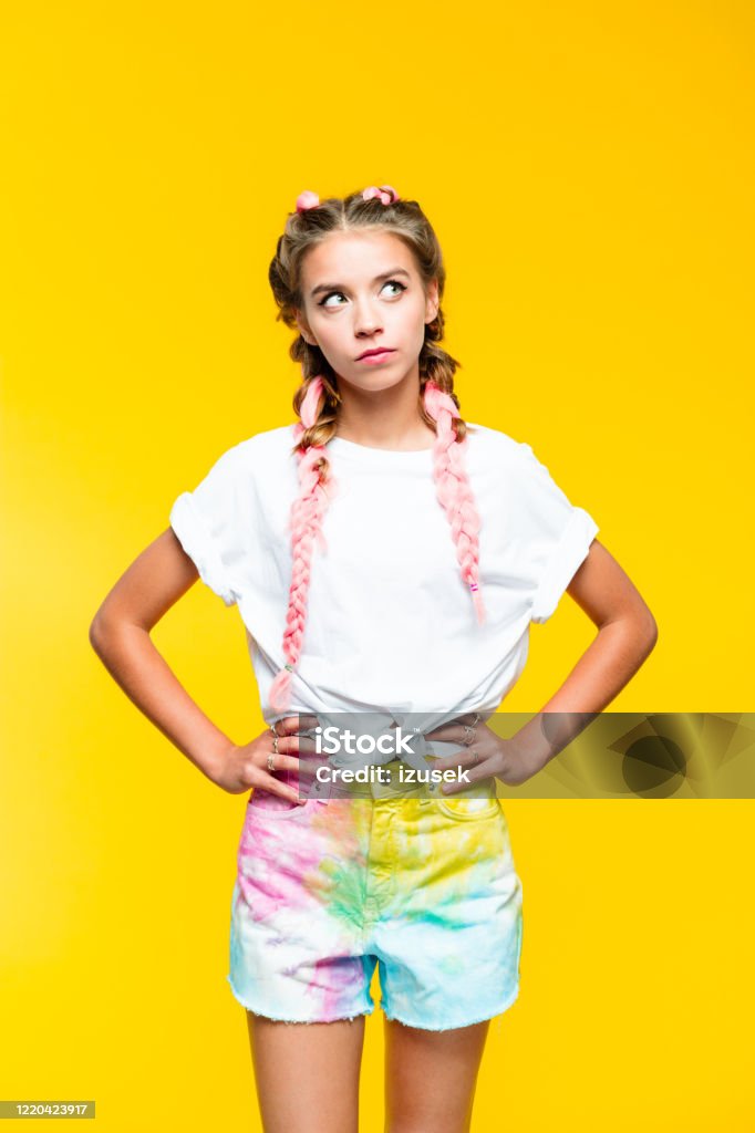 Teenage girl on yellow background looking away Displeased teenage girl with pink braids wearing white t-shirt and colorful shorts standing with hands on hip and looking away. Studio shot, yellow background. Party - Social Event Stock Photo