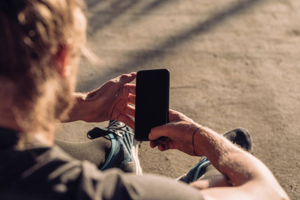 Over the Shoulder View of a Runner Using a Smartphone Outdoors (Blank Screen, Copy Space) Close up of a blond sportsman using his mobile phone outdoors. man looking at phone over shoulder stock pictures, royalty-free photos & images