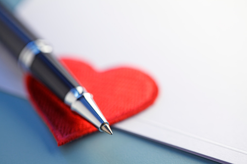 A ballpoint pen rests on a red heart shape and a blank sheet of paper stationery. Photographed with a very shallow depth of field.