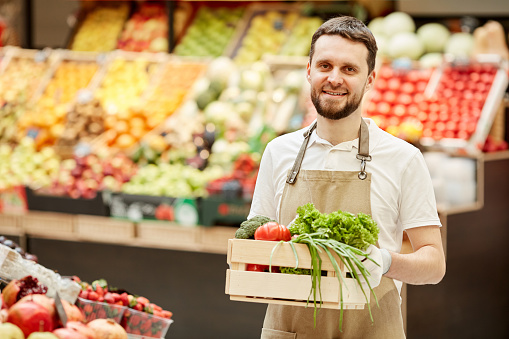 Waist up portrait of bearded man holding box of vegetables and smiling at camera while selling fresh produce at farmers market, copy space