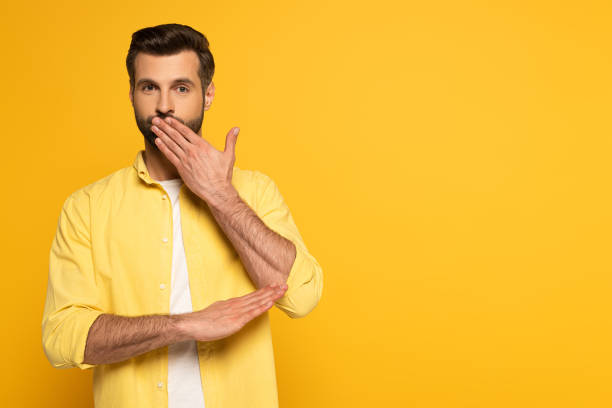 istock Handsome man covering mouth while using sign language on yellow background 1220435829