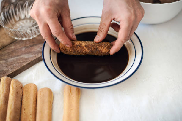 Quarantine cooking: a man is soaking ladyfinger-biscuits in coffee to prepare tiramisu Quarantine cooking: a man is soaking ladyfinger-biscuits in coffee to prepare tiramisu camel colored photos stock pictures, royalty-free photos & images