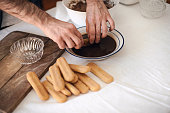 Quarantine cooking: a man is soaking ladyfinger-biscuits in coffee to prepare tiramisu