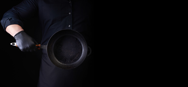 male cook in black uniform and latex gloves holds an empty round vintage black cast iron pan in front of him, low key, place for a restaurant to label its menu, schedule or price of dishes