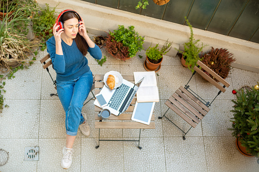 Aerial view of an attractive young woman working from home using a laptop, having a small breakfast, enjoying a beautiful day on her balcony