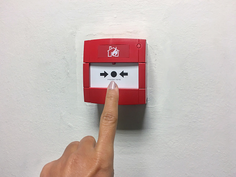 Close up shot of females finger pressing fire alarm button mounting on the white wall of apartment