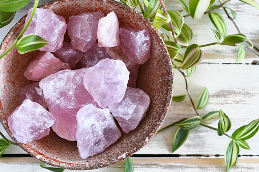 A close up image of a pottery bowl filled with raw rose quartz crystals and  a lush green plant on a white wooden table top.