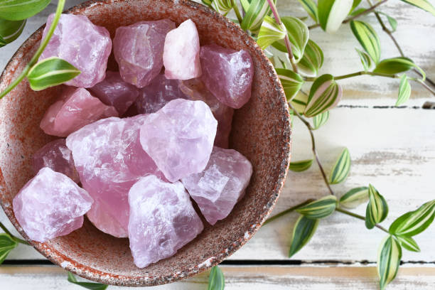 Rose Quartz Crystals A close up image of a pottery bowl filled with raw rose quartz crystals and  a lush green plant on a white wooden table top. crystal stock pictures, royalty-free photos & images