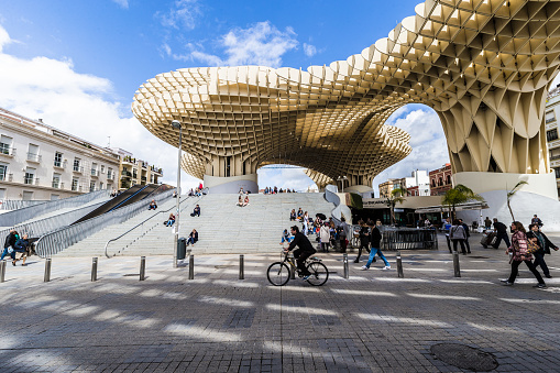 Seville, Spain - March 3, 2018 - La Encarnación Square in the old quarter of town at noon. The prominent wooden architecture is the Metropol Parasol by architect Jurgen Mayer. Both people of Seville and tourists are sitting on the steps, enjoying a beverage at the cafes and bars at ground level, or just passing by to cross the city.