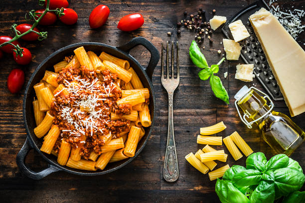 Italian food: rigatoni pasta with bologna sauce Italian food: rigatoni pasta with bologna sauce served in a cast iron pan shot from above on rustic wooden table. Parmesan cheese placed on a grater, fresh tomatoes, basil and an olive oil bottle complete the composition. High resolution 42Mp studio digital capture taken with Sony A7rII and Sony FE 90mm f2.8 macro G OSS lens rigatoni stock pictures, royalty-free photos & images