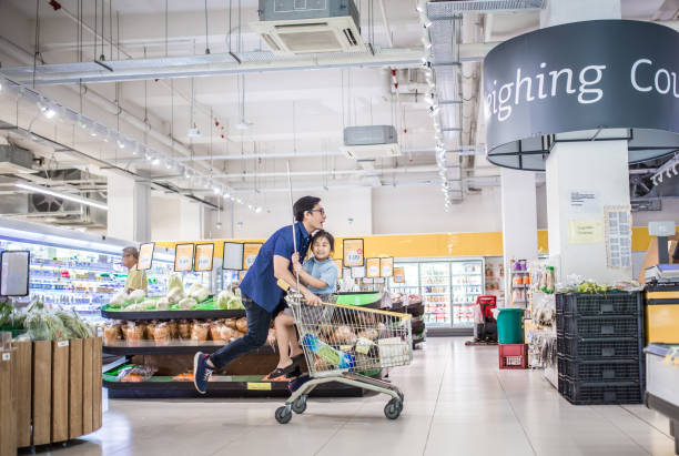 uma garota asiática chinesa sentada em um carrinho de compras sendo empurrada pelo pai. eles estão se divertindo no supermercado. - carrinho de puxar carrinho - fotografias e filmes do acervo