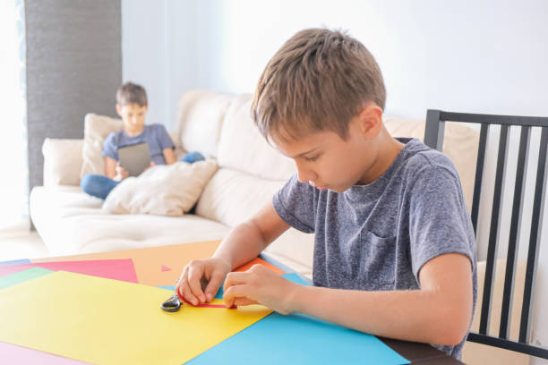 istock Education, online learning, entertiment at home. Boy making paper craft card with colored paper. Brother using tablet computer for online lesson at home 1220515478