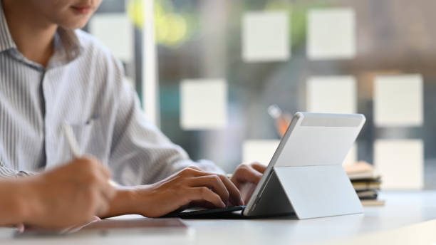 istock Cropped image of smart man typing on computer tablet with keyboard case while sitting at the white working desk with his colleague over modern office glass wall as background. 1220516495