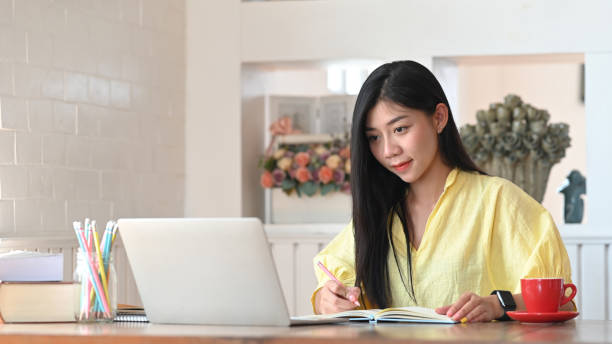 istock Photo of woman sitting and taking note while sitting in front her computer laptop that putting on wooden working desk and surrounded by coffee cup, pencil holder, potted plant and stack of books 1220516797
