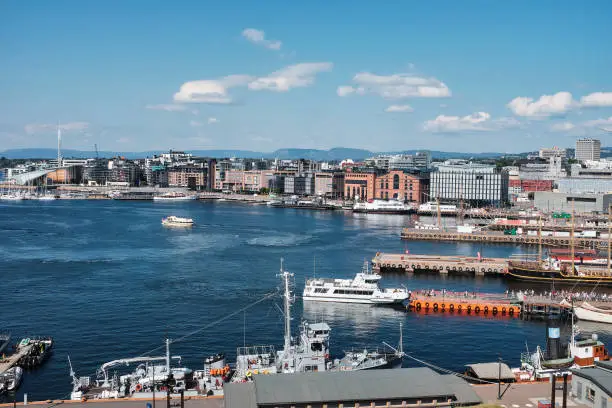 A wide view on the harbour of Oslo, Norway, on a warm summerday, with lots of modern architecture on the banks across. Tourists and inhabitants alike waiting in line on the pier for the ferries to get to the small Islands nearby, mostly for recreational purposes.