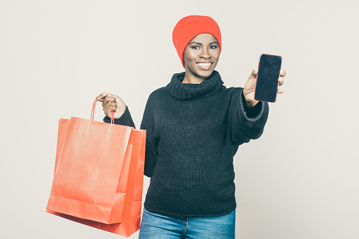 Smiling young woman showing smartphone with blank screen. Attractive African American lady holding smartphone and paper bags. Shopping, technology concept