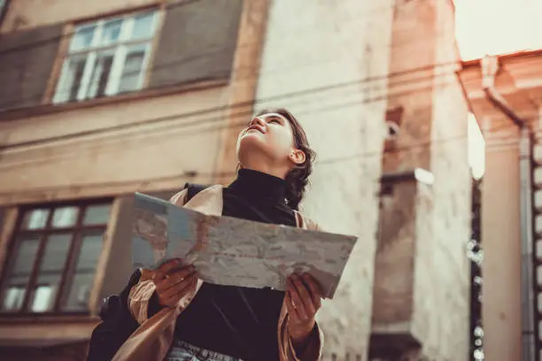 Photo of Bottom-view of pretty young female tourist studying a map, enjoying discovering a new city, looking excited at ancient architecture.