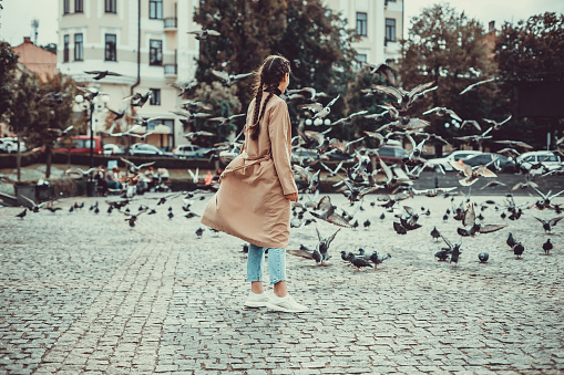 Concept of excitement. Photo of beautiful girl looking at pigeons on the square, flocking wings in cloudy autumn day.