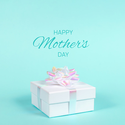 White gift box with holographic bow on pastel blue background. Shot at angle. Happy Mother's day square greeting card.