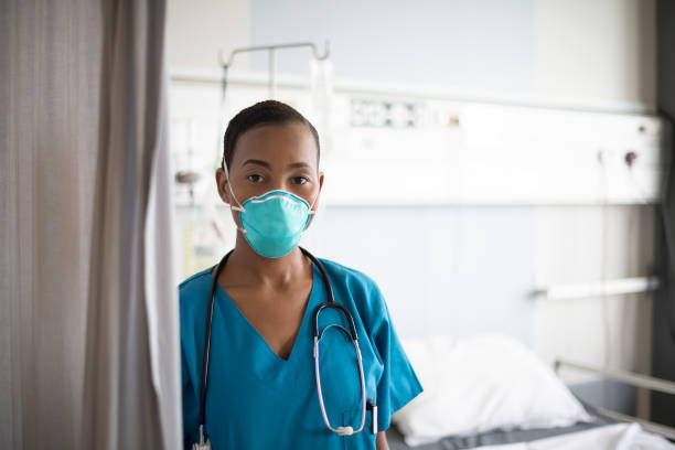 Indoor portrait of young African nurse wearing N95 face mask Close-up of serious African female nurse in mid 20s wearing blue scrubs, N95 face mask, and stethoscope around her neck standing in hospital room with empty bed. n95 face mask photos stock pictures, royalty-free photos & images