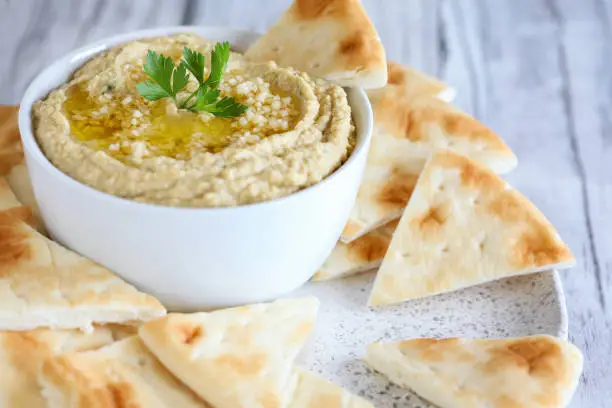 Vegan Hummus, made with chickpeas and tahini, with olive oil and garlic, garnished with parsley and served with pita bread over a white rustic table. Extreme shallow depth of field with background.