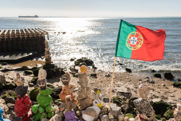 Lisbon, Portugal - February 12, 2019: Landscape view across sand beach on shore with flag, stone people structures and calm ocean beyond at the harbour in sunshine light of day.