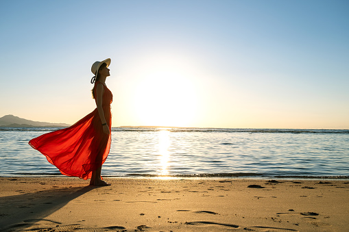 Young woman wearing long red dress and straw hat standing on sand beach at sea shore enjoying view of rising sun in early summer morning.