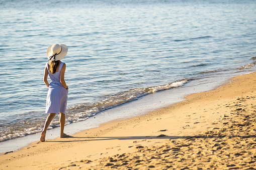 Young woman in straw hat and a dress walking alone on empty sand beach at sea shore. Lonely tourist girl looking at horizon over calm ocean surface on vacation trip.