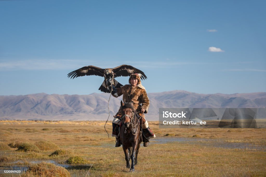 A young eagle hunter with his eagle and horse. A proud young kazakh eagle hunter posing with his golden eagle on horseback on the backdrop of blue sky. Ulgii, Mongolia. Kazakhstan Stock Photo