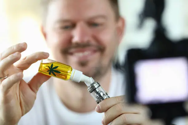 Close-up view of smiling middle-aged man dropping cannabinoid oil into special container. Happy male filming process on video camera. Beginner blogger making experiment
