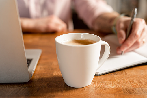 A man is taking notes on a note pad. In front of him is a fresh cup of hot coffee. Focus on foreground.