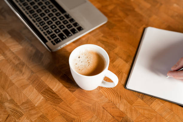 A cup of fresh coffee next to a note pad and a laptop on a wooden table.