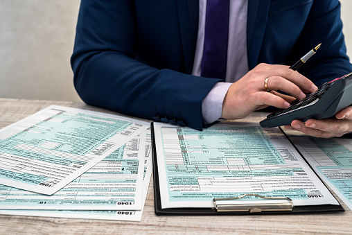 A man in a business suit writes a 1040 tax form in the office. Male hands fill on paper with calculator at workplace. Accounting concept