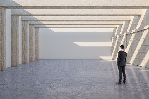 Businessman standing in minimalistic gallery interior with empty wall and wooden columns. Mock up,