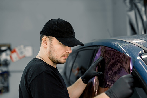 Professional car service worker wearing black cap and t-shirt, tinting a car window with tinted foil or film in auto workshop. Tinting of car windows.