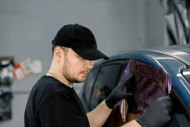 Photo of Professional car service worker wearing black cap and t-shirt, tinting a car window with tinted foil or film in auto workshop. Tinting of car windows